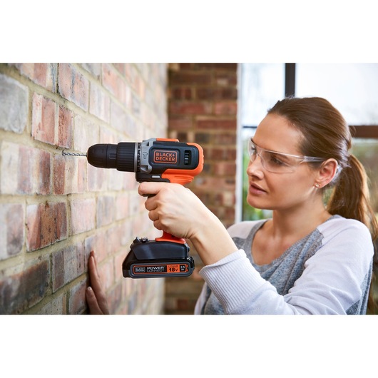 A person using a BLACK+DECKER cordless drill to make a hole in a brick wall. The drill is orange and black with visible branding and an 18V lithium battery. The person's face is blurred for privacy.