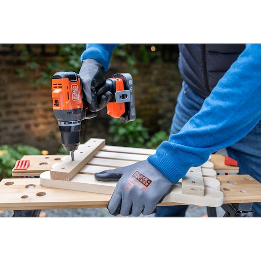 Person uses BLACK+DECKER cordless drill on wood, wearing branded gloves, outdoors on workbench.