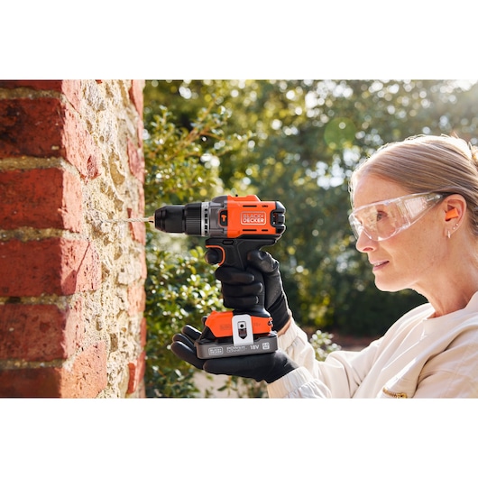 Person using BLACK+DECKER cordless drill to drill into a brick and stone wall outdoors, wearing safety glasses.