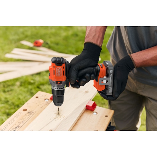 BLACK+DECKER cordless drill being used to drill a hole in a wooden plank outdoors by a person wearing gloves.