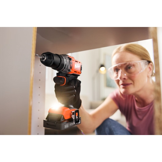 Person using a BLACK+DECKER cordless drill to assemble furniture, wearing protective glasses and gloves.
