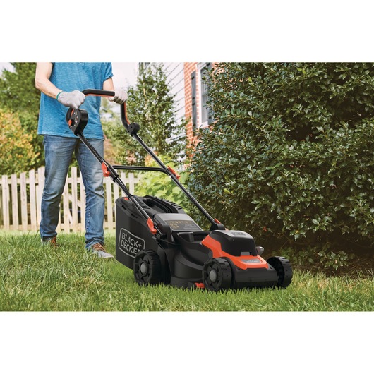 A person mowing the lawn with a BLACK+DECKER cordless lawn mower in a residential garden. The mower is black and orange, and the person is wearing gloves and jeans.