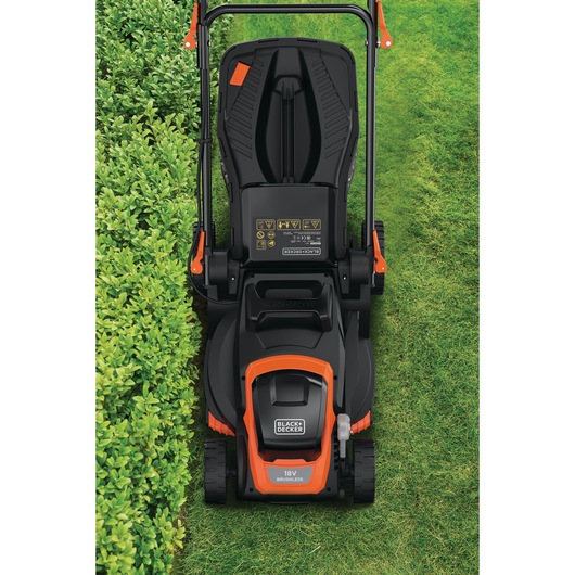Top view of a BLACK+DECKER 18V cordless lawn mower positioned on a lawn, showing the handle, control panel, and part of a trimmed hedge on the left side.