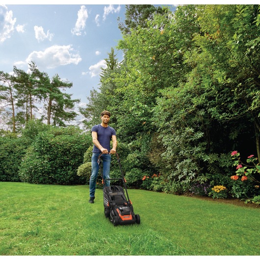 A person with a blurred face is mowing a lawn with a BLACK+DECKER lawn mower in a garden surrounded by green bushes and trees under a blue sky.
