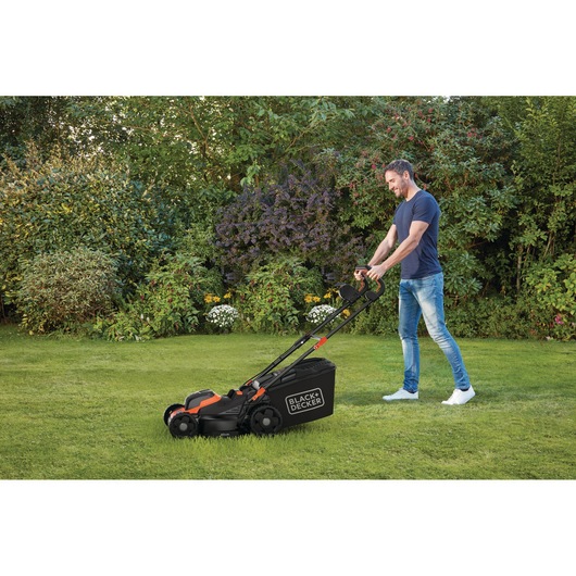 A person is using a BLACK+DECKER lawn mower on a grassy lawn in a garden, surrounded by bushes and flowers.