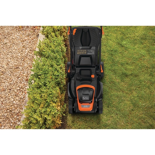 Top view of a BLACK+DECKER lawn mower cutting grass near a hedge and a gravel path.