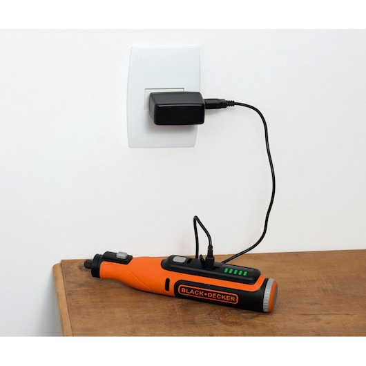 A BLACK+DECKER orange and black electric tool is shown charging on a wooden surface, connected to a power outlet through a black charger and cable.