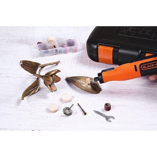 A BLACK+DECKER rotary tool is being used to polish a decorative metal flower. Various rotary tool accessories and a wrench are laid out on the table. In the background, there is a black carrying case and a small organizer box with additional tool bits.