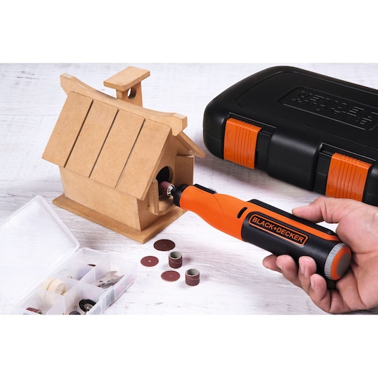 A person uses a BLACK+DECKER rotary tool to sand the entrance of a wooden birdhouse. The scene includes a black and orange carrying case and an open box with various sanding and grinding accessories on a light wooden surface.