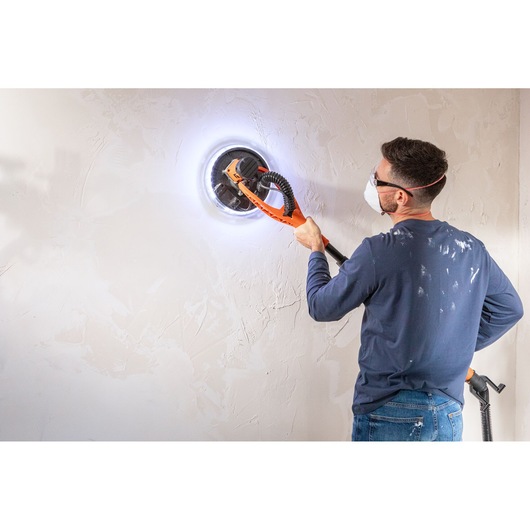 Person uses BLACK+DECKER wall sander with LED light on a white wall, wearing safety glasses and mask.