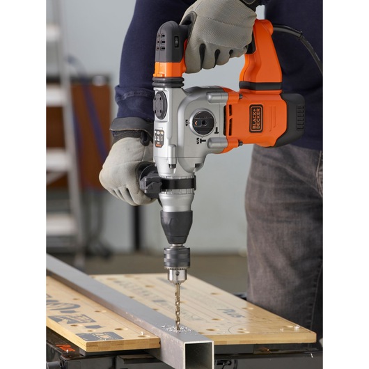 BLACKDECKER BEHS03K hammer drill in use, drilling metal on a workbench, with user wearing gloves.