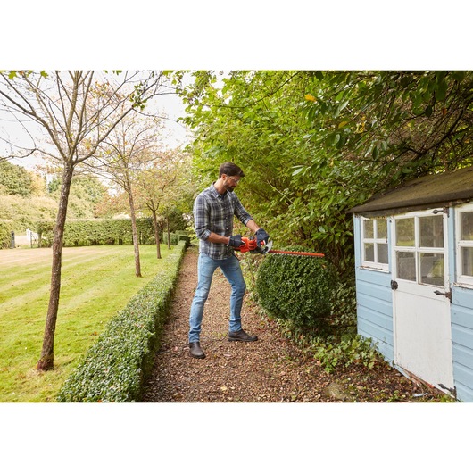 Man trimming garden hedge with BLACKDECKER BEHT201 hedge trimmer near blue shed in a yard.