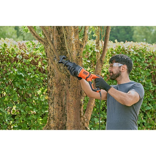 A person wearing gloves is using a BLACK+DECKER BES301 reciprocating saw to cut a tree branch outdoors. The saw is orange and black, and the scene features greenery in the background.