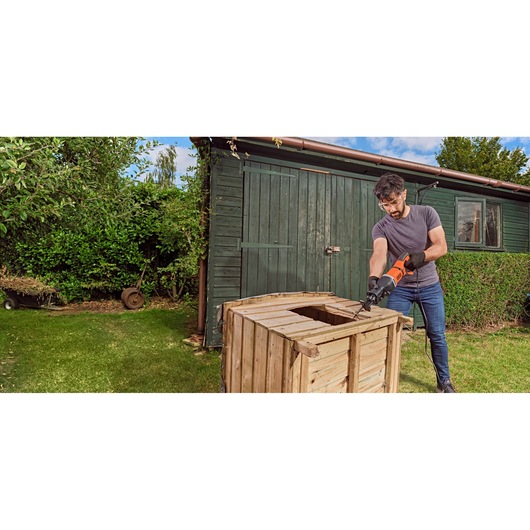 A person uses a BLACK+DECKER reciprocating saw (model BES301) to cut a wooden crate outdoors near a green shed. The individual is wearing gloves and working on the lawn.