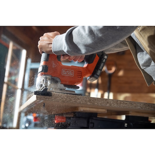 Photo of BLACKDECKER BLCJS78E1 jigsaw cutting wood panel, close-up of hands operating orange cordless tool.