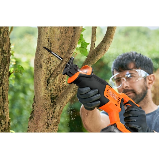 BLACK+DECKER BLCR78E1 reciprocating saw cutting tree branch outdoors, shown in use with gloved hands.