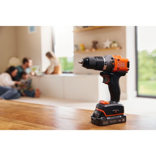 Photo of BLACK+DECKER cordless drill BLD683D1 with black and orange design on wooden table, blurred family in background.
