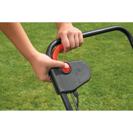 Close-up of a person's hands operating the handle and start button of a BLACK+DECKER lawn scarifier, with green grass in the background.
