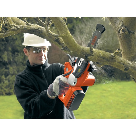 A person wearing work gloves and a hard hat is using a BLACK+DECKER cordless chainsaw to cut a thick tree branch outdoors. The face of the person is blurred for privacy.