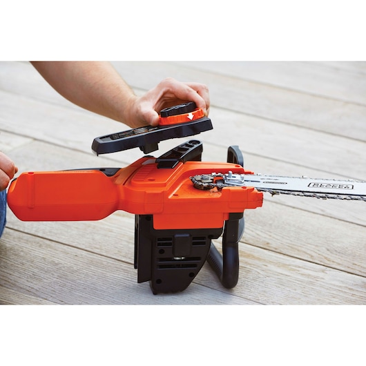 A person is performing maintenance on an orange and black cordless chainsaw, with the cover of the chain mechanism being lifted to show the internal parts. The chainsaw is placed on a wooden surface.