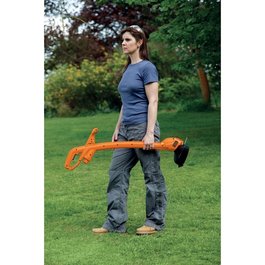 A person standing outdoors on grass, holding an orange BLACK+DECKER GL310 grass trimmer. The person's face is blurred, and they are wearing casual clothing.