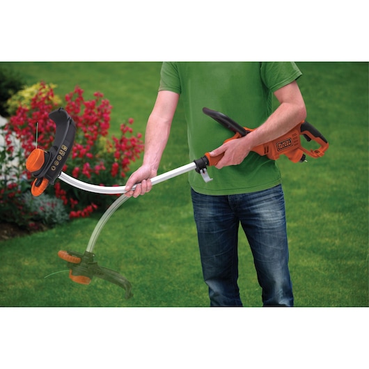 A person holding a BLACK+DECKER GL7033 grass trimmer while standing on a lawn. The orange and black electric grass trimmer is shown in use, with flowers and greenery in the background.