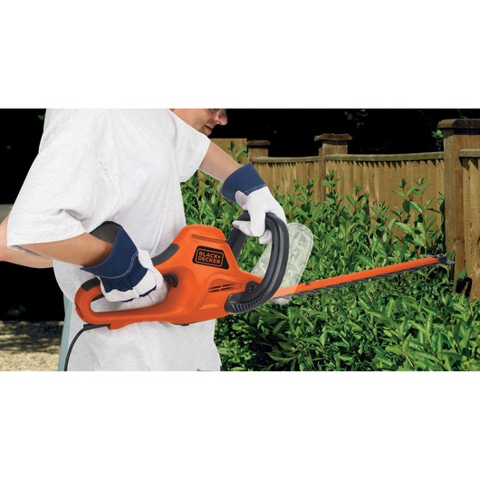 A person wearing gloves uses an orange BLACK+DECKER electric hedge trimmer to trim green bushes along a wooden fence outdoors.