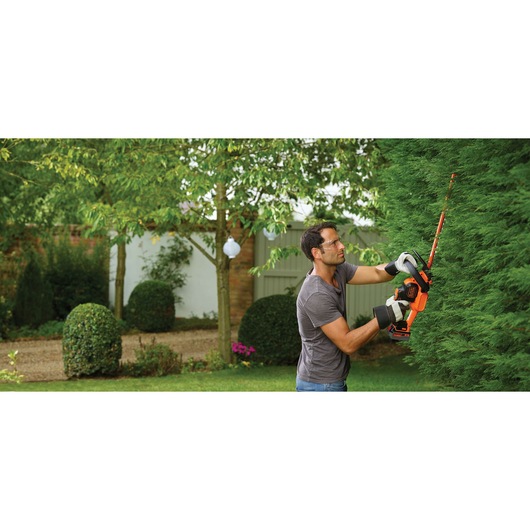 A person using a BLACK+DECKER cordless hedge trimmer (model GTC18504PC) to trim a tall green hedge in a garden. The person is wearing gloves and a grey shirt, and the garden features trimmed bushes and trees in the background.