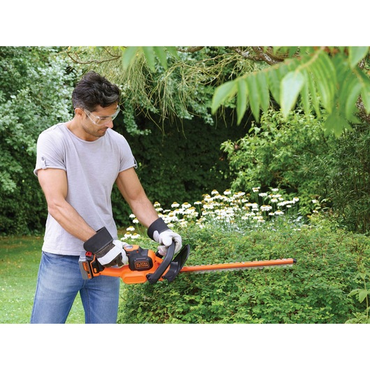 A person using a BLACK+DECKER cordless hedge trimmer (model GTC18504PC) to trim bushes in a garden, wearing gloves and a grey t-shirt with their face blurred for privacy.