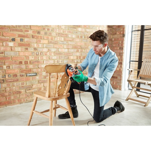 A person wearing a green glove is using a BLACK+DECKER electric sander to sand a wooden chair in a room with a brick wall. The sander is orange and black.