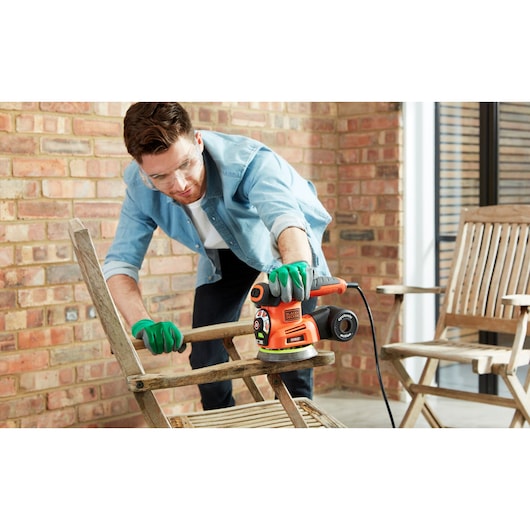 A person wearing green gloves uses an orange BLACK+DECKER KA280LSA2 power tool to sand a wooden chair in an outdoor patio setting. Other wooden chairs and a brick wall are visible in the background.