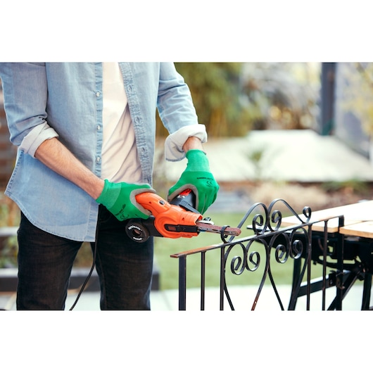 A person wearing green gloves uses an orange BLACK+DECKER electric tool to sand or grind a metal fence outdoors.