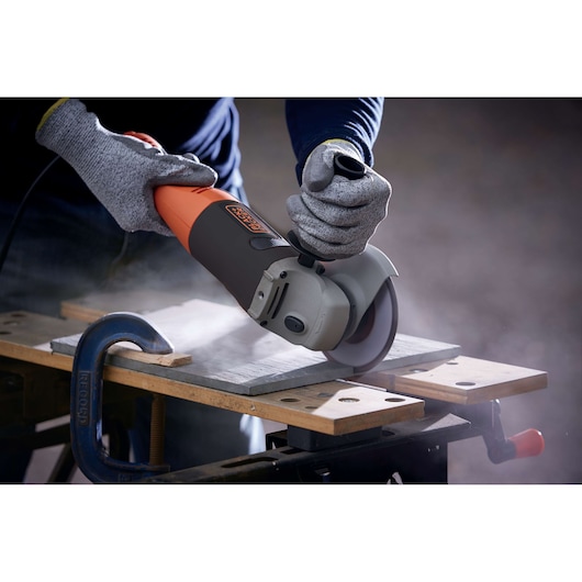 A person wearing protective gloves uses a BLACK+DECKER angle grinder to cut a piece of material on a workbench, with visible dust being generated.