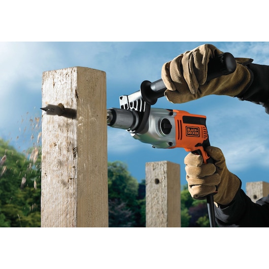 A person wearing gloves uses a BLACK+DECKER electric drill to bore a hole into a wooden post outdoors.