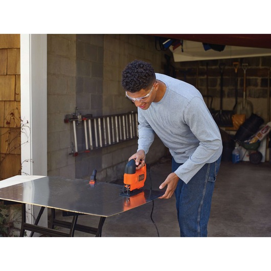 Person using a BLACK+DECKER KS701E jigsaw to cut a sheet of material on a work table in a garage. The person's face is blurred for privacy.
