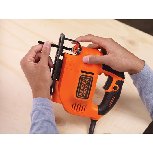 A person is installing a saw blade into an orange and black BLACK+DECKER jigsaw on a light wooden surface.