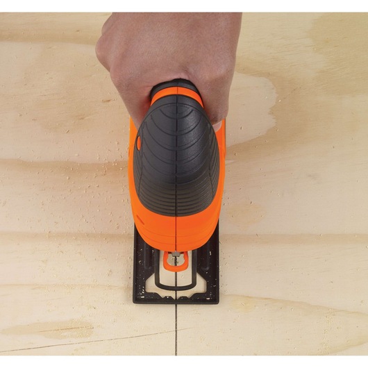 A person's hand holding an orange and black BLACK+DECKER jigsaw, cutting a straight line in a piece of wood.