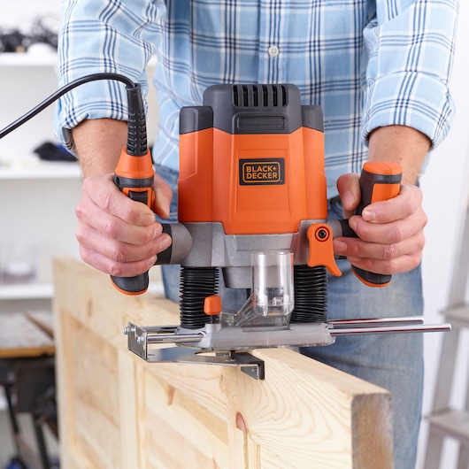 Photo of BLACK+DECKER KW1200EKA wood router in use on a plank, front view, orange and black tool with clear base.