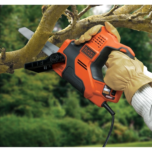 A person wearing gloves uses a BLACK+DECKER electric reciprocating saw to cut a tree branch outdoors.