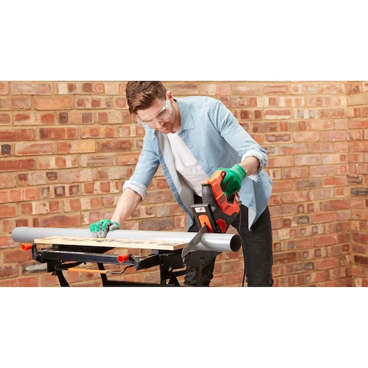 A person wearing green gloves is using a BLACK+DECKER RS890K electric saw to cut a piece of wood placed on a workbench. The scene is set against a brick wall background.