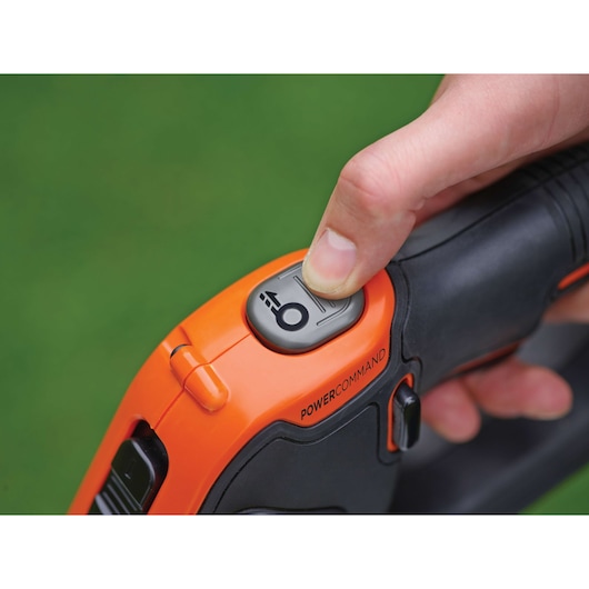 Close-up of a hand pressing the POWERCOMMAND button on an orange and black BLACK+DECKER gardening tool, with a green background.
