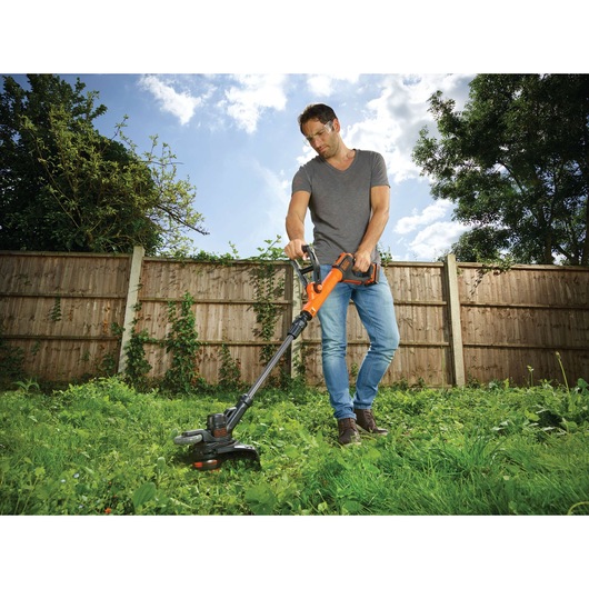 A person using an orange and black BLACK+DECKER cordless grass trimmer on a lawn in a backyard, with a wooden fence and trees in the background.